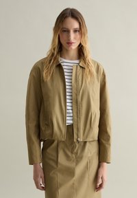 Chaqueta corta de color verde oliva con cierre de cremallera, bolsillos laterales y cuello. Usada sobre una blusa a rayas blanca y azul marino y una falda a juego.