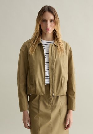 Giacca corta verde oliva con zip frontale, tasche laterali e colletto. Indossata su un top a strisce bianche e blu e una gonna abbinata.