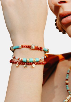 Trois bracelets empilés avec des perles rouges, des accents turquoise, du matériel doré et des charms décoratifs. Couleurs et textures distinctes.