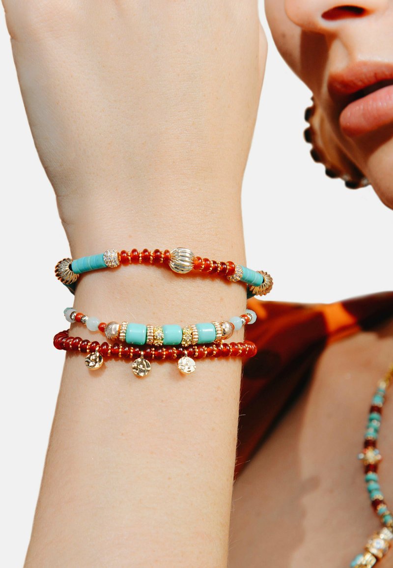 Trois bracelets empilés avec des perles rouges, des accents turquoise, du matériel doré et des charms décoratifs. Couleurs et textures distinctes.