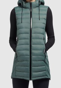 Kobieta w bezrękawniku w kolorze turkusowym, pikowanym, z kapturem, zapinanym na zamek błyskawiczny i bocznymi kieszeniami, nosi czarną bluzkę z długim rękawem oraz legginsy.