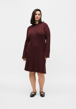 Femme portant une robe en maille côtelée à manches longues rouge foncé avec des chaussures Mary Jane noires, debout devant un fond gris clair uni.