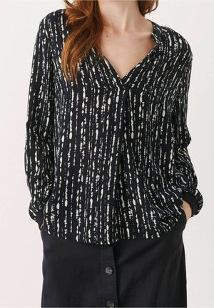 Woman wearing a black long-sleeve blouse with white vertical abstract patterns and a black button-front skirt, hands in pockets.