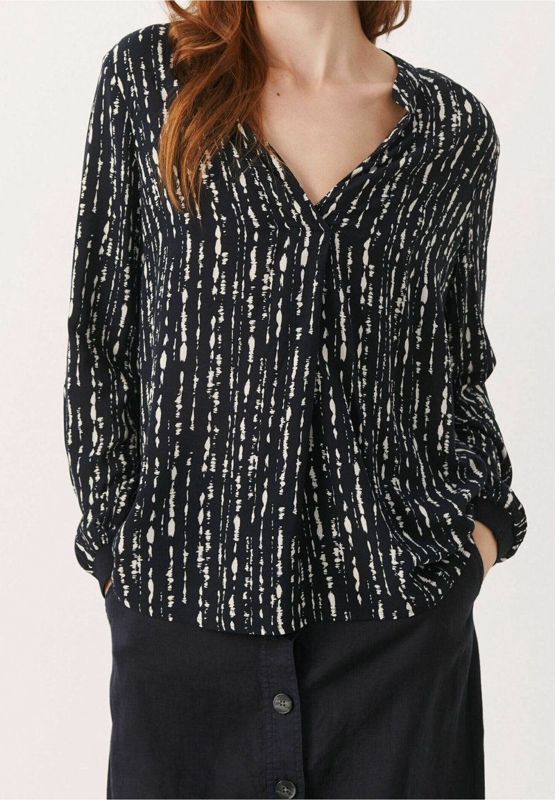 Femme portant un chemisier noir à manches longues avec des motifs abstraits verticaux blancs et une jupe noire boutonnée devant, mains dans les poches.