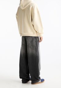 Beige mikina s kapucí prostorného střihu, v kombinaci s volnými, vybledlými černými džíny se zadními kapsami. Obuví jsou modré sandály.