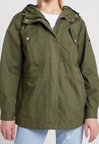 Olive green jacket with a hood, two front pockets, adjustable waistband, and a zippered closure. Made of lightweight fabric.