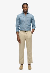 Lichtblauwe chambray shirt, beige lichtgewicht broek, beige instap schoenen. Langgerekte vorm, ontwerp met kraag en taille met trekkoord.