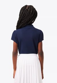 Flicka med långt flätat hår som har på sig en marinblå polo och en vit veckad kjol, vänd bort från kameran.