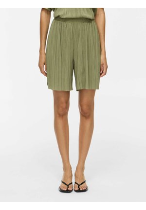 Femme portant un short plissé vert olive et un haut assorti, debout avec des tongs noires sur un fond blanc uni.