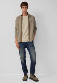 Beige gebreide cardigan met een gestructureerd patroon, bruine rits, lichtbeige shirt eronder, gecombineerd met donkerblauwe jeans en bruine veterschoenen.
