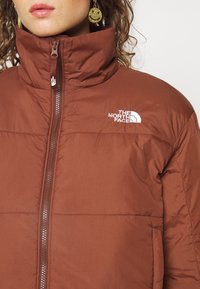 Braune Daunenjacke mit hohem Kragen, durchgehendem Reißverschluss und weißen Logodetails. Sie verfügt über eine glatte Textur und eine isolierte Bauweise.