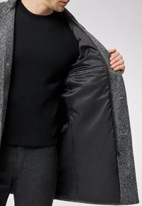 Grau karierter Mantel mit glatter schwarzer Innenseite, ausgestattet mit einer Brusttasche und detaillierter Naht entlang der Kanten.