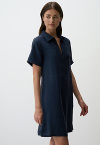 Robe chemise bleu marine avec un col, des manches courtes et une fermeture éclair à l'avant. Texture lisse et forme en A avec des fentes latérales.