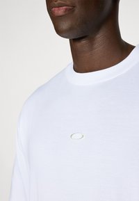 Hvid bomulds t-shirt med rund hals, der har et lille, broderet logo i sølv i midten. Glat tekstur, korte ærmer, afslappet pasform.