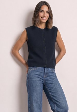 Femme souriante portant un haut noir sans manches boutonné et un jean bleu, debout les mains sur les hanches devant un mur clair uni.