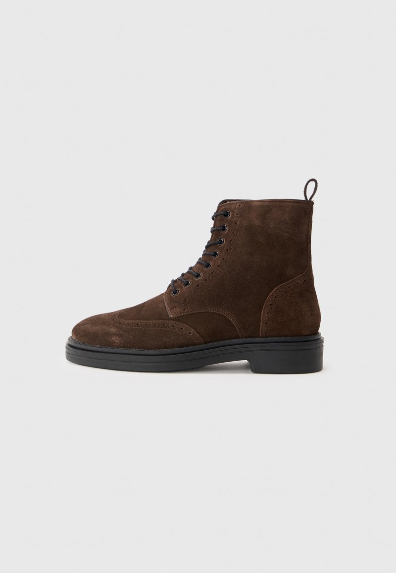 Botas de tobillo de ante marrón con puntera redondeada, cordones negros, costuras decorativas y suela de goma texturizada. Asa en el talón para ponerlas con facilidad.