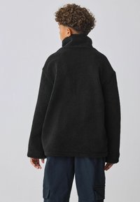 Zwarte fleece pull met hoge kraag, oversized pasvorm en een gestructureerd oppervlak. Lange mouwen en ontspannen ontwerp. Draagt met donkere cargobroek.