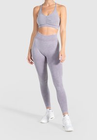 Sujetador deportivo lila con detalle fruncido y leggings de talle alto a juego. El material parece suave con una textura lisa. Zapatillas blancas completan el look.