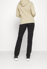 Beige hoodie med snörning i huvan, kombinerad med svarta byxor och ljusa sportskor, sedd bakifrån. Texturerad bomullstyg.