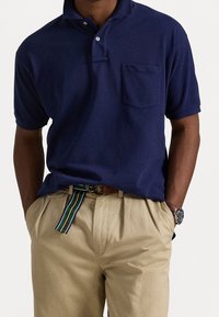 Námořnická modrá polo košile s krátkými rukávy a límečkem, se svrchní kapsou, doplněná o světle béžové kalhoty a páskem s multibarevným proužkem.