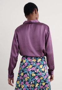 Camicia lunga in satin viola con colletto a punta, dotata di polsini con bottoni, abbinata a una gonna con fantasia fiorita su uno sfondo scuro.