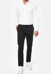 Ein Mann trägt ein weißes Langarm-Hemd mit Knöpfen, dunkle Slim-Fit-Hosen und weiße Sneakers und steht mit einer Hand in der Tasche.