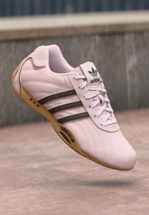 Jasnoróżowy sneaker Adidas z brązowymi paskami i gumową podeszwą, pokazany w powietrzu na rozmytym tle zewnętrznego chodnika.