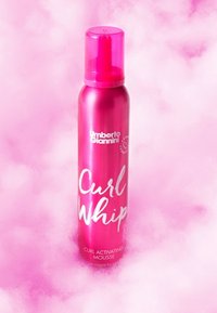 Pink metallic aerosol canister labeled "Curl Whip" with white text, featuring a pink cap. The background is soft pink with a fluffy texture.