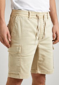 Bermudas color beige fabricadas en algodón, con una cinturilla ajustable con cordón, bolsillos laterales y detalles de dobladillo doblado. Tejido texturizado con un acabado suave.