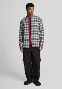 Geruite button-up shirt in grijs, wit en marineblauw over een bordeauxrode crew neck t-shirt, gecombineerd met zwarte cargo broeken en donkerbruine slippers.