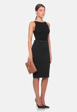 Donna con un vestito nero senza maniche con una cintura sottile, che tiene una pochette marrone e indossa scarpe nere con tacco alto.