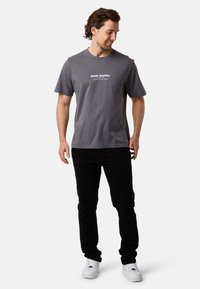 Harmaan puuvillapaitaan, jossa on lyhyet hihat ja pyöreä pääntie, on painettu valkoisella "Slade Studios" -teksti. Paita on yhdistetty mustiin housuihin ja valkoisiin lenkkareihin.