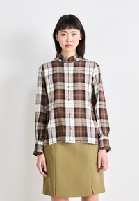 Knoop-up geruite blouse in bruin, crème en blauwe tinten. Lange mouwen met ruches aan de mouwen. Gecombineerd met een groene A-lijn rok met uitsnijdingen.