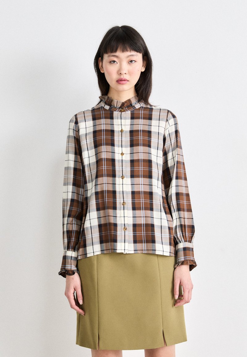 Knoop-up geruite blouse in bruin, crème en blauwe tinten. Lange mouwen met ruches aan de mouwen. Gecombineerd met een groene A-lijn rok met uitsnijdingen.