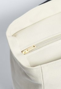 Plátěná taška s krémovým povrchem, se zlatým zipem a strukturovaným tvarem. Interiér je hladký a bez podšívky.