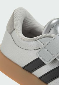 Close-up of a light gray sneaker with black stripes, gray mesh lining, a velcro strap, and a gum rubber sole.