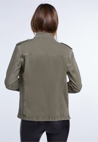 Chaqueta de mezclilla verde oliva, ajustada, con epaulettes en los hombros, costuras visibles y un dobladillo recto. La vista trasera muestra una textura suave y un diseño entallado.