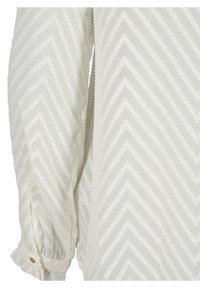 Bílá texturovaná halenka se vzorem cikcak, s nařasenými rukávy zdobenými knoflíky. Lehká látka s hladkým povrchem.
