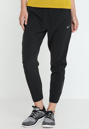 Person, der bærer sorte Nike joggebukser og grå og sorte Nike løbesko, står mod en lys baggrund.