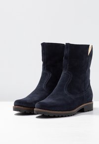 Námořnické semišové kotníkové boty s nízkým blokem, detailní šití po stranách a kontrastní hnědý panel na zadní straně. Odolná gumová podešev pro stabilitu.