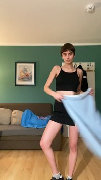 Crna crop top majica i mini suknja na osobi koja drži svijetloplavu tkaninu. Pozadina uključuje smeđi kauč i zelenu zidnu površinu. Pod je drveni.