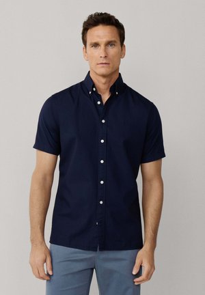Homme portant une chemise à manches courtes bleu marine avec boutons et un pantalon bleu clair, debout devant un fond gris clair uni.