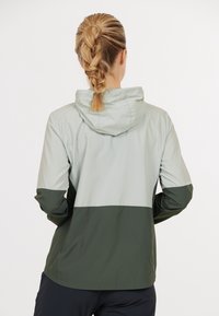 Veste imperméable légère bicolore avec une partie inférieure verte et une partie supérieure gris clair, dotée d'une capuche et d'une finition lisse et texturée.
