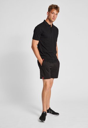 Schwarzes Poloshirt mit kurzen Ärmeln und Kragen, kombiniert mit schwarzen Shorts. Die Shorts verfügen über Seitentaschen und ein kleines rotes Etikett.