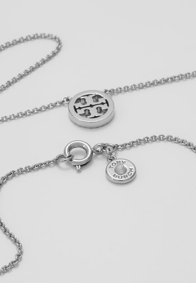 Colier din metal argintiu cu un pandantiv circular, având un design cu logo decupat și un mic pandantiv gravat cu "TORY BURCH."