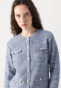 Knitted blue cardigan featuring a textured fabric, round neckline, six gold-button closures, and two chest pockets.