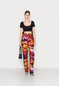 Top preto oversized combinado com calças vibrantes de várias cores com padrões de olhos abstratos. Os acessórios incluem uma pequena bolsa preta e sandálias de salto.