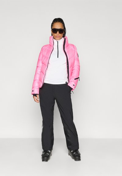 Jachetă puf din material roz, cu guler înalt și fermoar frontal, asortată cu un top alb și pantaloni de schi negri. Botinele de schi negre completează ținuta.