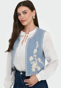 Ljusblå väst med vit blomsterbroderi och detaljerad kantning, bärs över en vit texturerad blus. Mörka jeans fulländar outfiten.