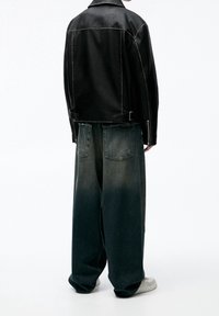 Black leather jacket with contrasting stitching, paired with wide-leg, faded blue jeans and white sneakers. View from the back.